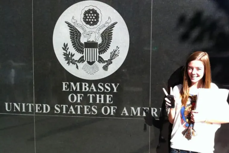 Chica sonriente sosteniendo documentos frente al sello oficial de la Embajada de Estados Unidos