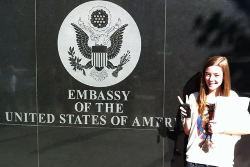 Chica sonriente sosteniendo documentos frente al sello oficial de la Embajada de Estados Unidos