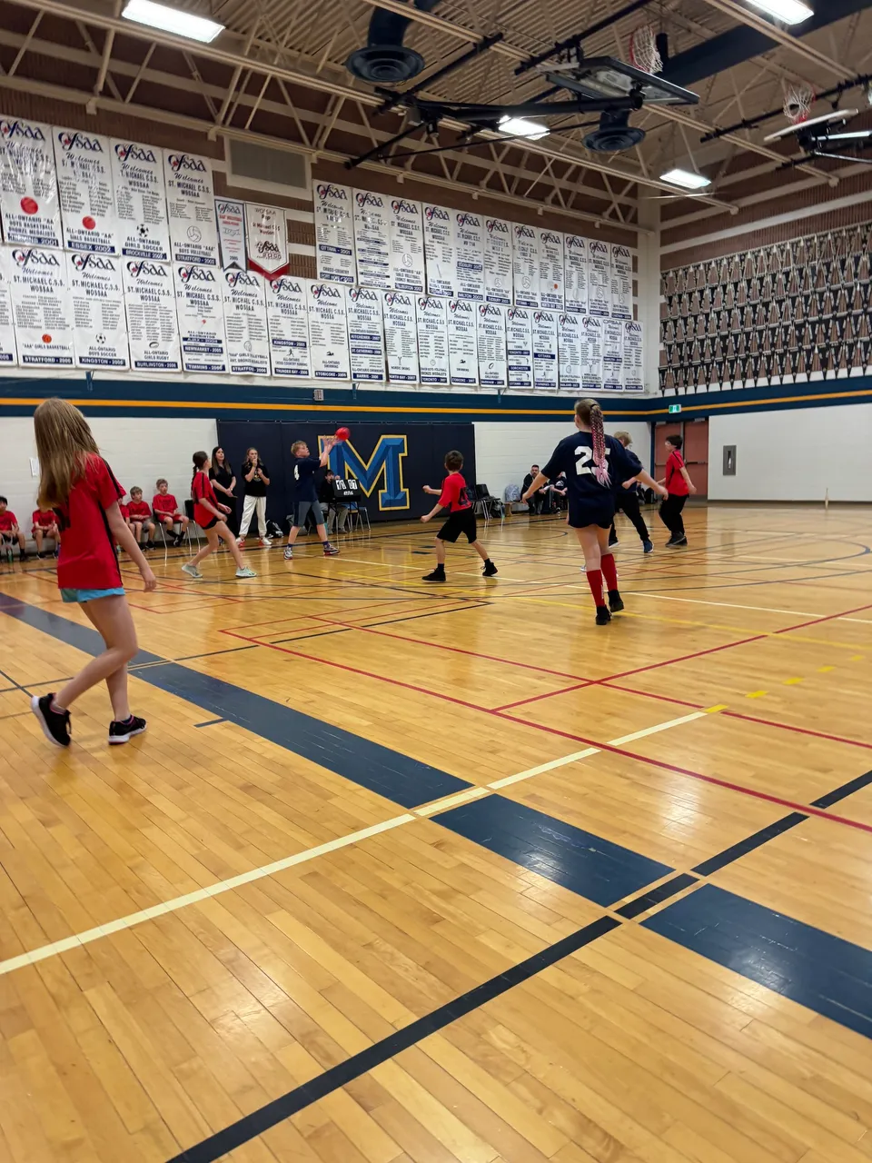 Studenti giocano a basket sul campo in legno con il grande logo M presso il Huron-Perth Catholic District School Board.
