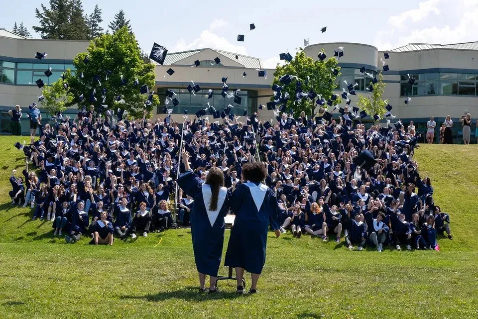 Un gruppo di diplomati della Salmon Arm Secondary in toga nera si trova su un prato davanti a un edificio e festeggia il diploma.
