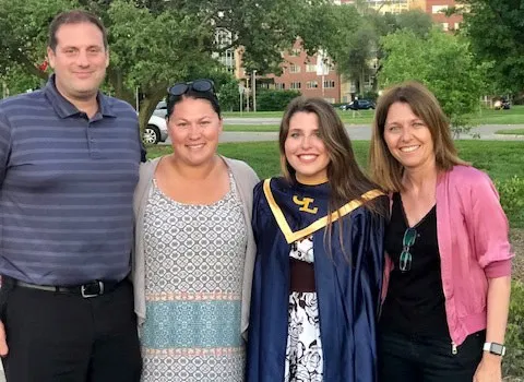 Una joven con túnica azul de graduación sonríe junto a tres adultos en un parque