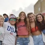 Cinco chicas jóvenes sonriendo juntas al aire libre, una vistiendo camiseta blanca con letras USA