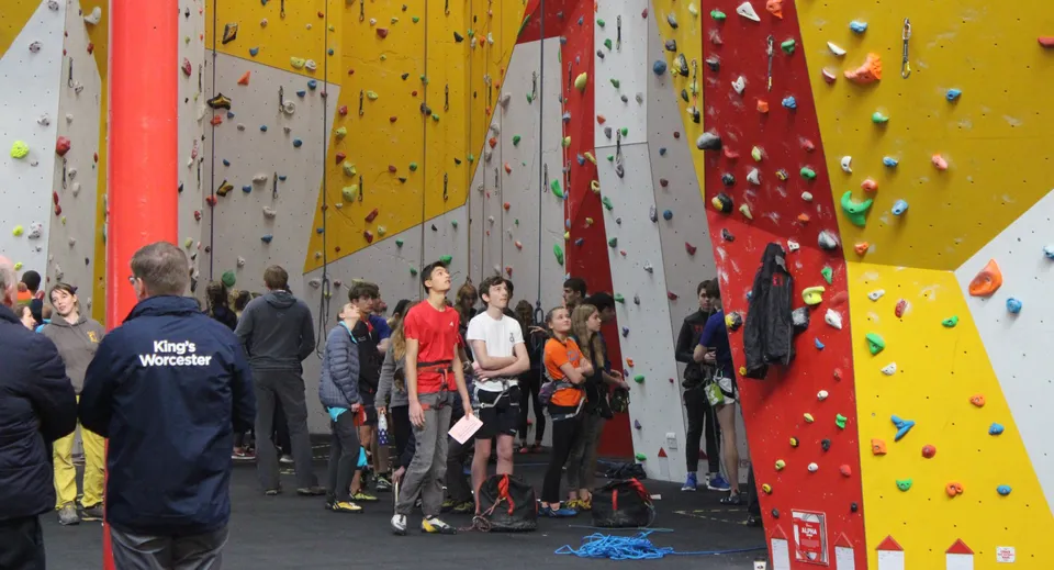 Studenti della Bredon School con attrezzatura da arrampicata si trovano davanti alla parete colorata nella palestra da arrampicata.