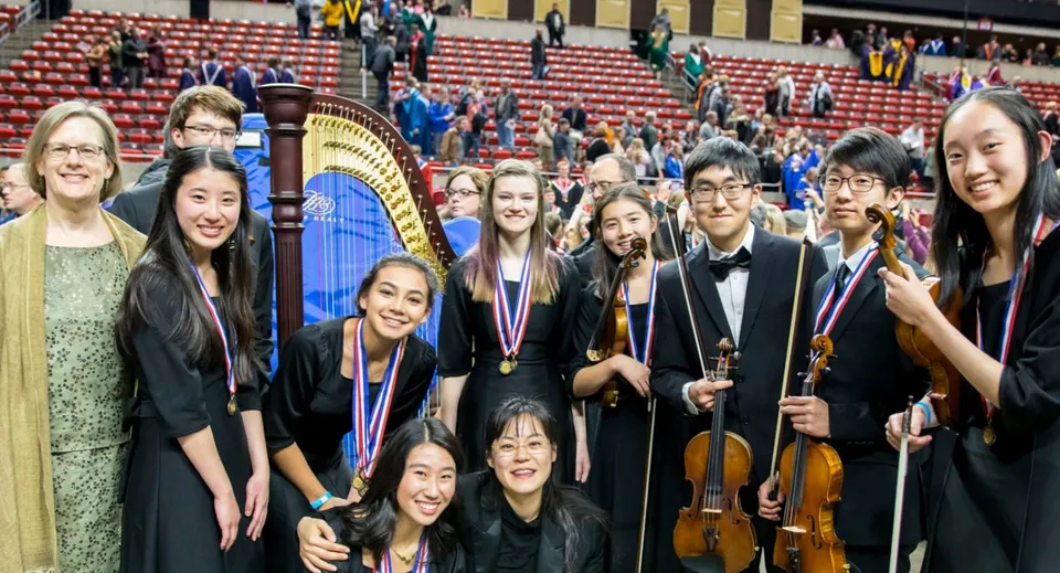 Un gruppo di giovani musiciste e musicisti in posa sul palco del Cranbrook Education Campus davanti a poltroncine rosse e a una grande arpa.