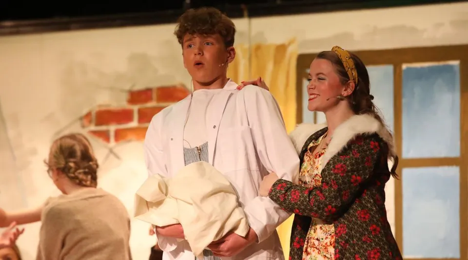 Due persone si trovano sul palco della Royal School Armagh davanti a una parete di mattoni con oggetti di scena sullo sfondo.