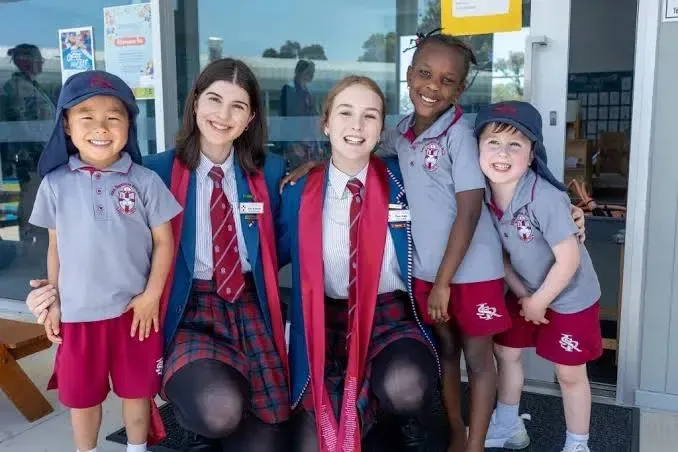 Cinco estudiantes de distintas edades de la John Septimus Roe Anglican Community School sonríen en uniforme frente a un edificio.