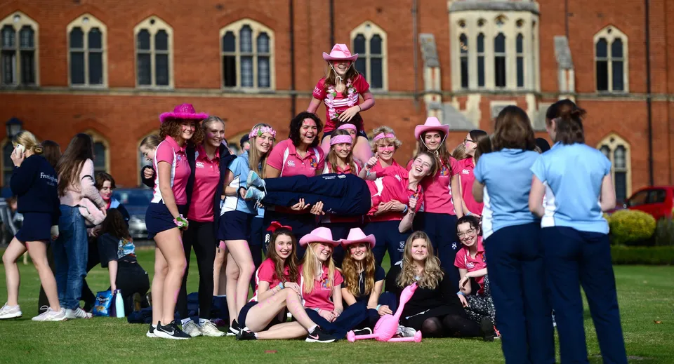 Un gruppo di studentesse del Framlingham College in outfit rosa si trova su un prato davanti a un edificio in mattoni rossi con finestre ad arco.