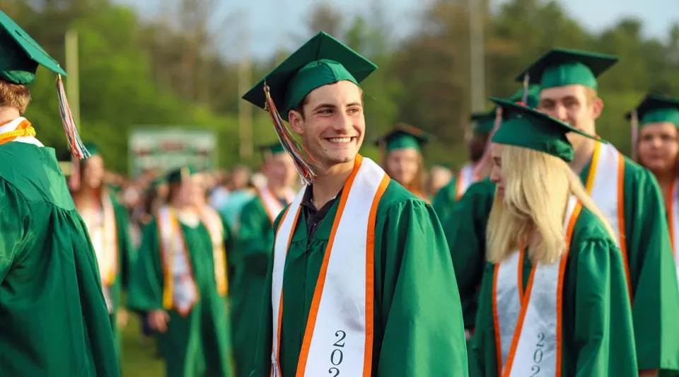 Un gruppo di diplomati e diplomate del Hopkinton Public School District indossa toghe verdi e arancioni davanti a uno sfondo boschivo.