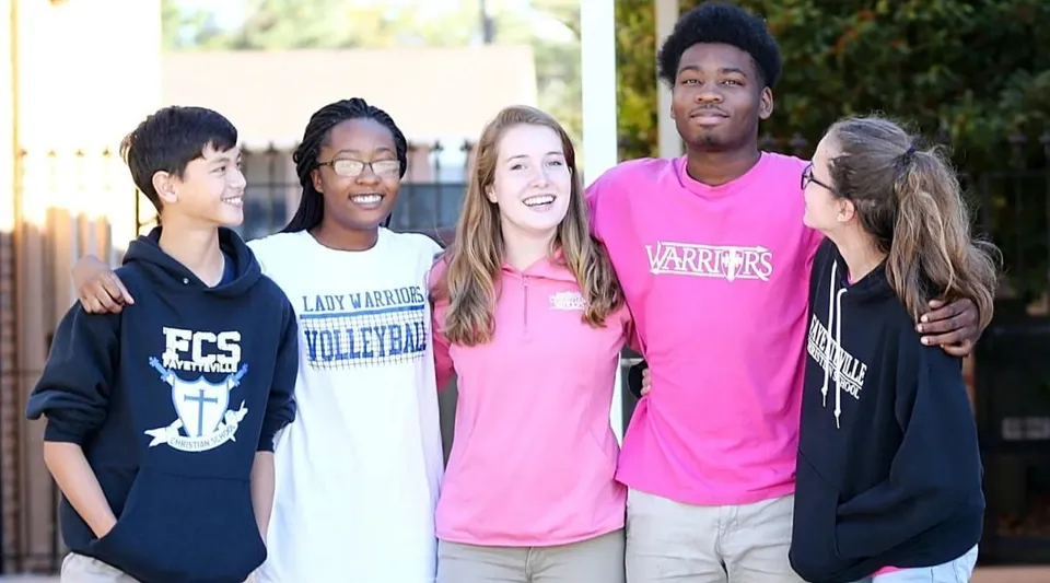 Un gruppo di studenti della Fayetteville Christian School si trova all'aperto con divise sportive davanti a una recinzione con alberi.