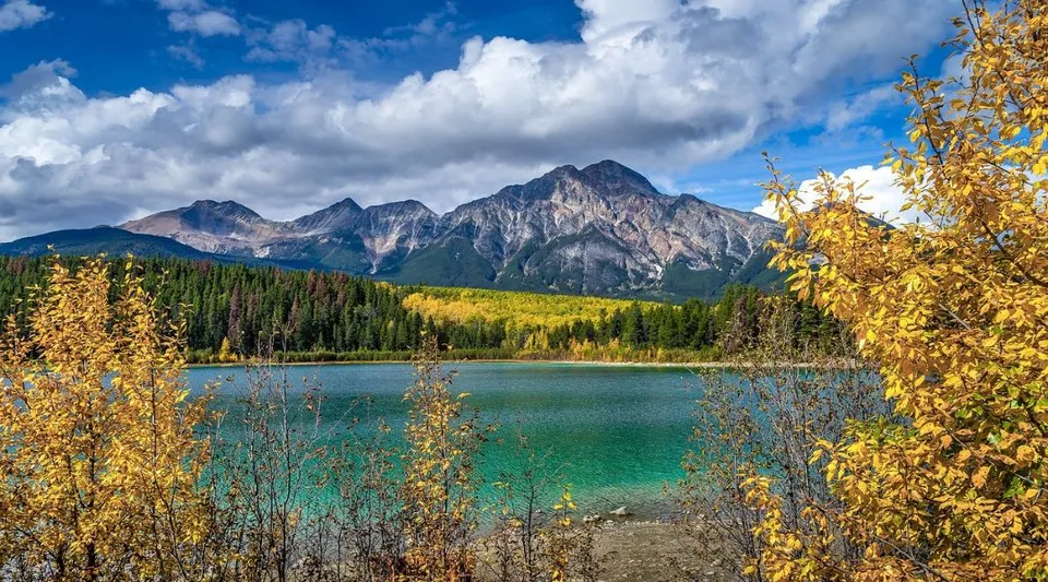 Un lago turchese circondato da alberi con foglie autunnali dorate davanti a una catena montuosa.