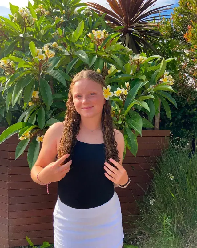 Studentessa sorridente con un fiore di frangipani tra i capelli davanti a piante tropicali