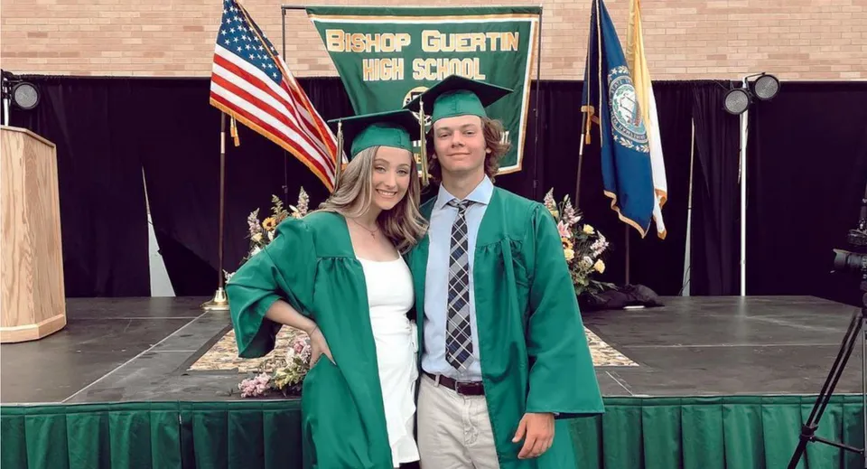 Due studenti della Bishop Guertin High School in abiti da diploma davanti a uno sfondo con la bandiera americana e quella della scuola.