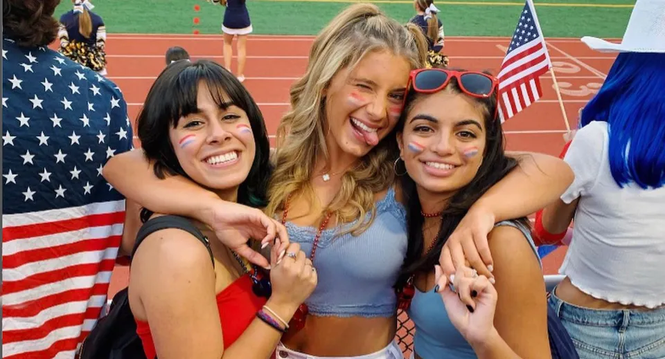 Tre studentesse del Las Virgenes Unified School District si abbracciano sorridenti davanti a una bandiera americana su un campo sportivo.