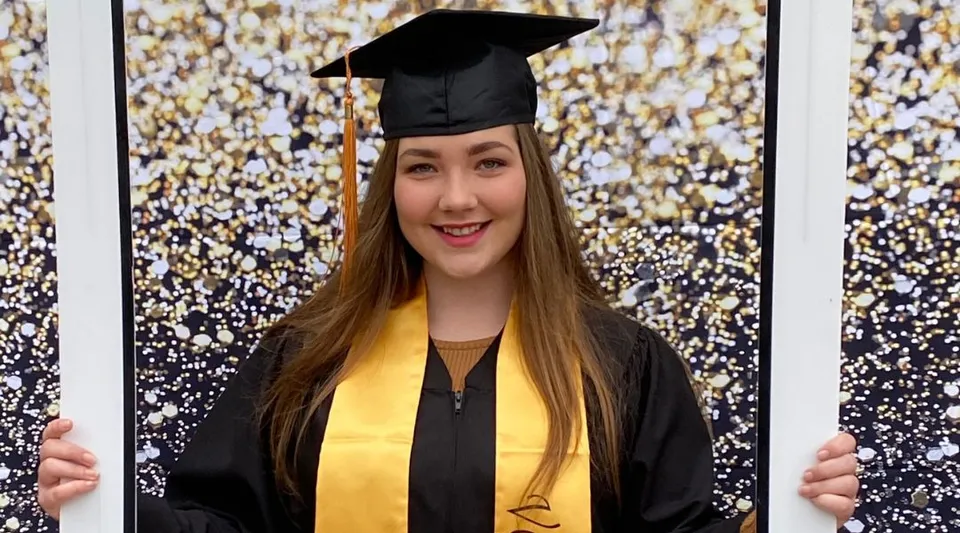 Una giovane donna in toga da diploma è in piedi davanti a uno sfondo scintillante nel campus della St. Michael's Regional High.