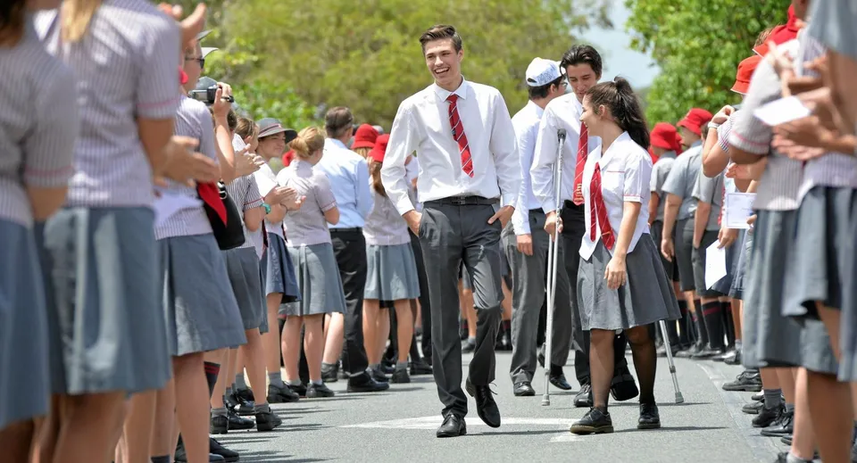 Un gruppo di studenti della Whitsunday Anglican School in abito formale cammina lungo un viale circondato da spettatori durante una cerimonia di diploma.