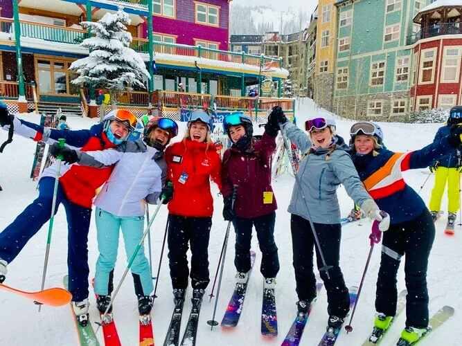 Un grupo de estudiantes de la Salmon Arm Secondary está de pie con equipo de esquí de colores en un paisaje nevado frente a edificios y un árbol cubierto de nieve.