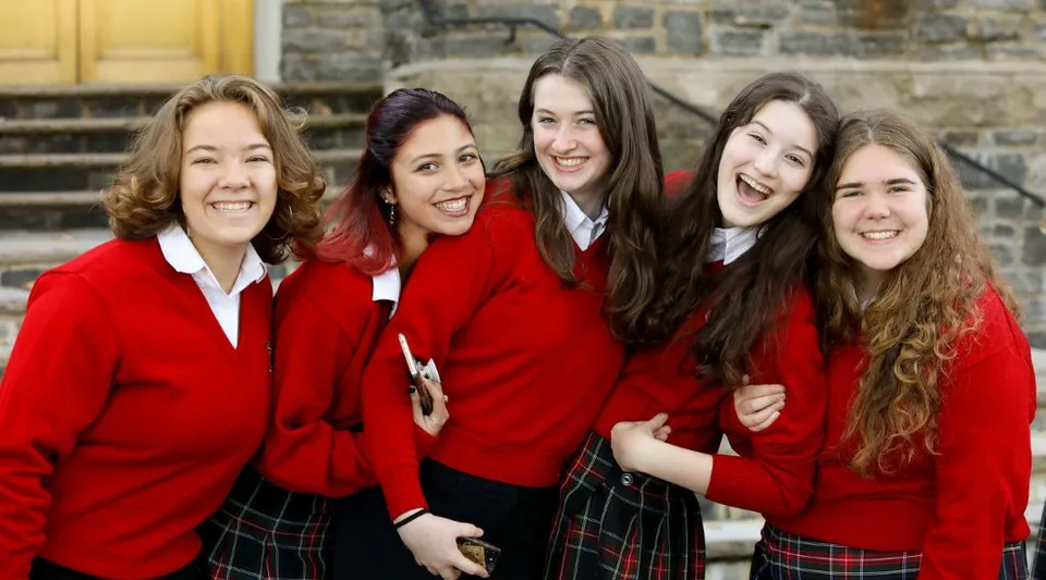 Cinque studentesse in divisa scolastica rossa posano davanti a un edificio in pietra nel campus della Academy of the New Church.