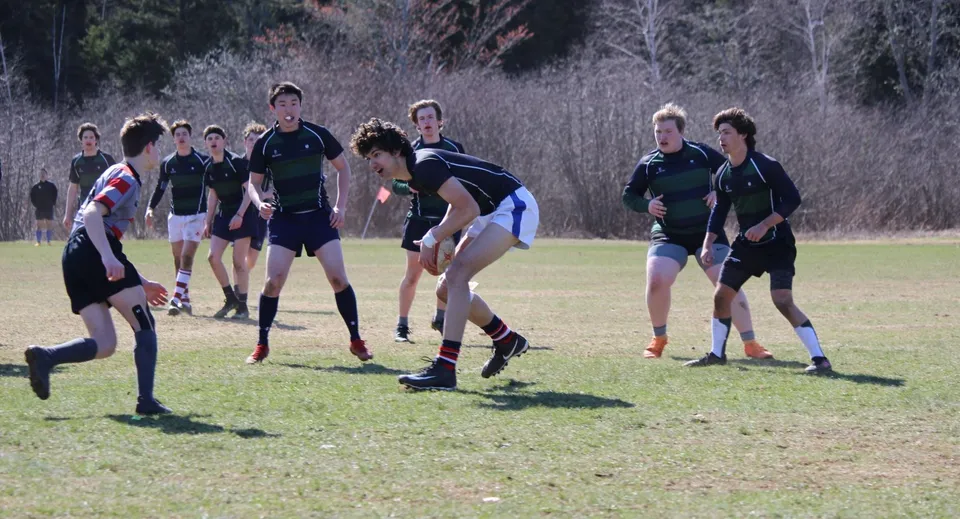Studenti della Rothesay Netherwood School giocano a rugby su un campo d'erba davanti a un bosco.