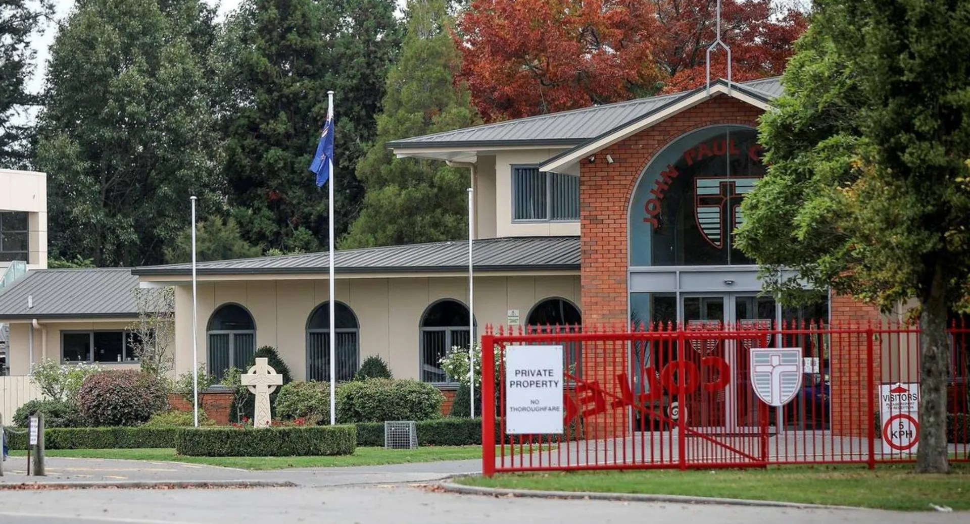 L'edificio in mattoni con il portone rosso del John Paul College è circondato da alberi e aree verdi.