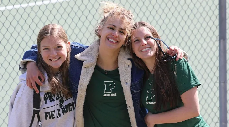 Tre studentesse della Proctor Academy si abbracciano ridendo davanti a una rete metallica.