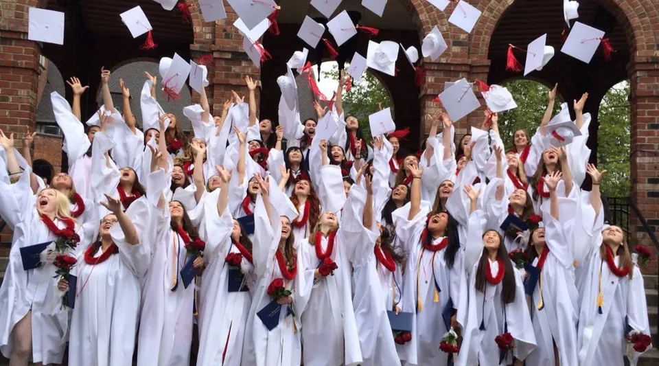 Un gruppo di studenti in tuniche bianche tiene bandiere rosse e bianche nel campus delle Annie Wright Schools.