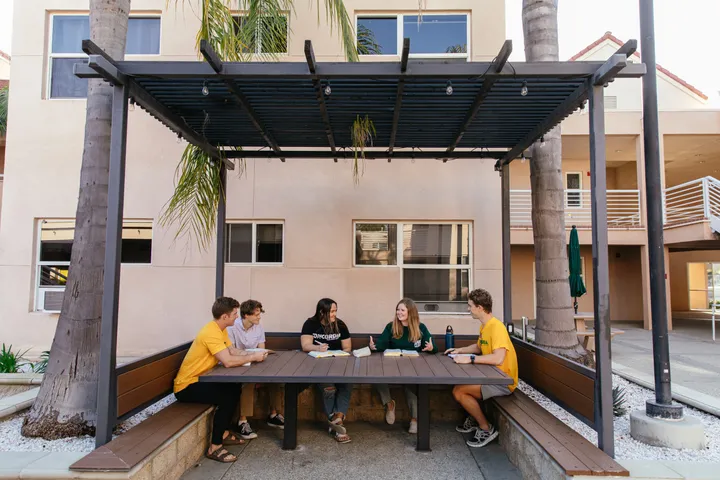 Zona exterior del campus con pérgola de madera, mesa larga y estudiantes sentados a la sombra