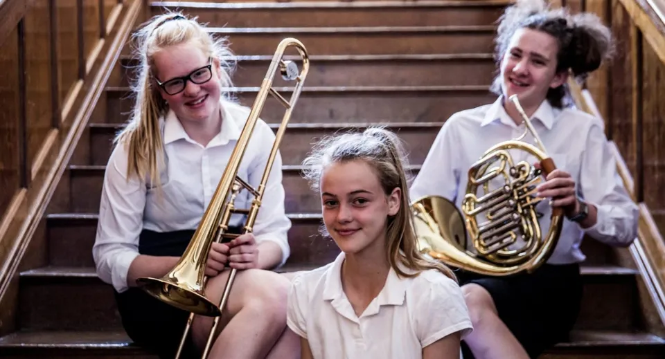 Tre studentesse della Queenswood School sono in piedi su una scala di legno davanti a uno sfondo in legno con un trombone e un corno francese.