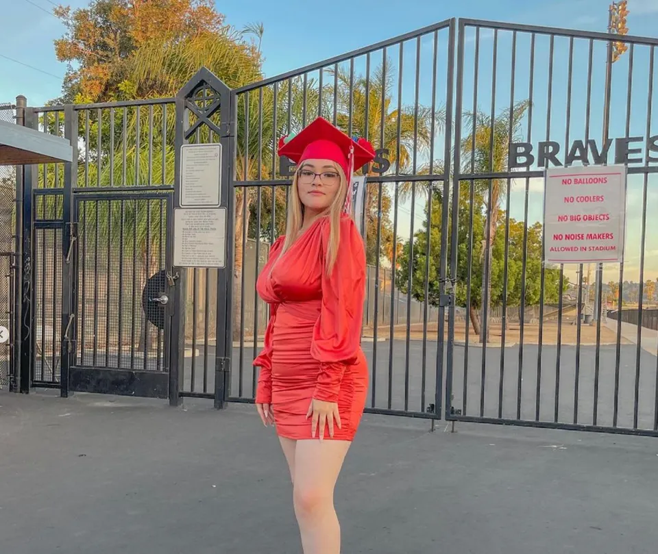 Una donna in un vestito rosso in piedi davanti a una recinzione metallica con la scritta Braves nel campus della El Cajon Valley High School.