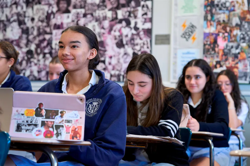 Un gruppo di studenti è seduto in un'aula della Notre Dame High School con un collage di immagini e poster sullo sfondo.