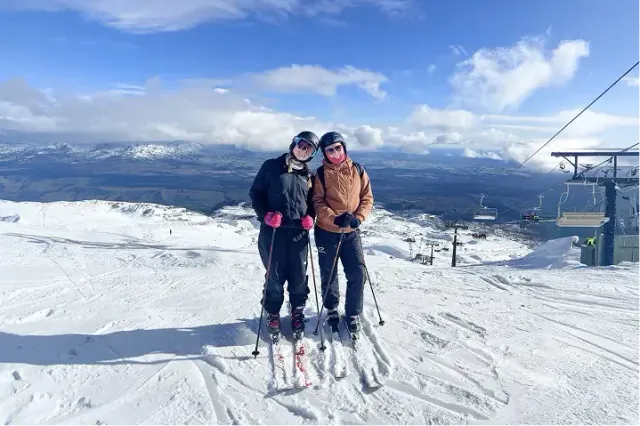 Due amiche con sci e caschi in cima a una montagna innevata sotto il cielo azzurro, in Nuova Zelanda