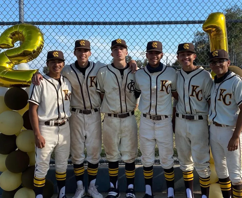 Un gruppo di giovani giocatori di baseball si trova davanti a una recinzione in rete metallica nel campus della John F. Kennedy High School.