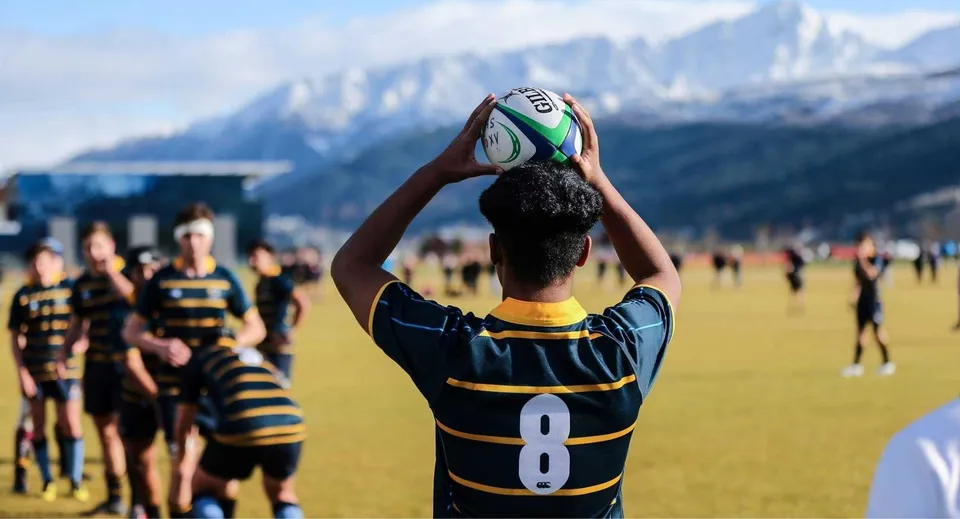 Un giocatore di rugby in divisa nera e gialla afferra la palla sul campo da gioco della Wakatipu High School con montagne innevate sullo sfondo.