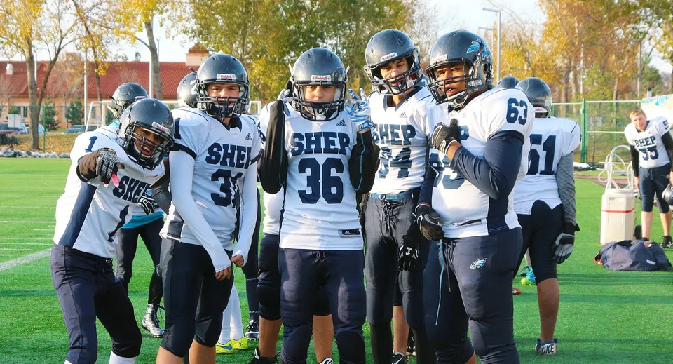 Un gruppo di giocatori di calcio della Ross Sheppard High School si trova insieme su un campo d'erba con alberi ed edifici sullo sfondo.