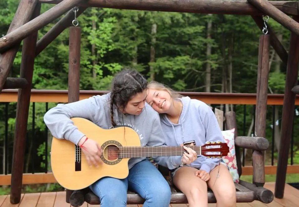 Due studentesse della Thornton Academy siedono su una panchina di legno nel bosco, una suona la chitarra e l'altra ascolta.