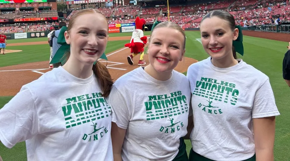 Tre studentesse della Metro East Lutheran High School con t-shirt bianche posano sorridenti in uno stadio di baseball.