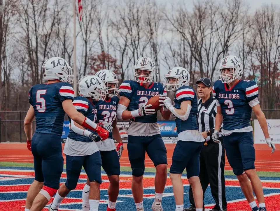 Un gruppo di giocatori di football sta sul campo della Liberty Christian Academy davanti a uno sfondo alberato.