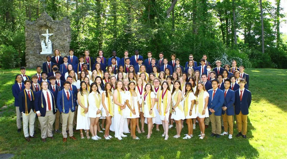 Un gruppo di studentesse e studenti della Marianapolis Preparatory School è in abbigliamento formale davanti a un monumento a forma di croce all'aperto.