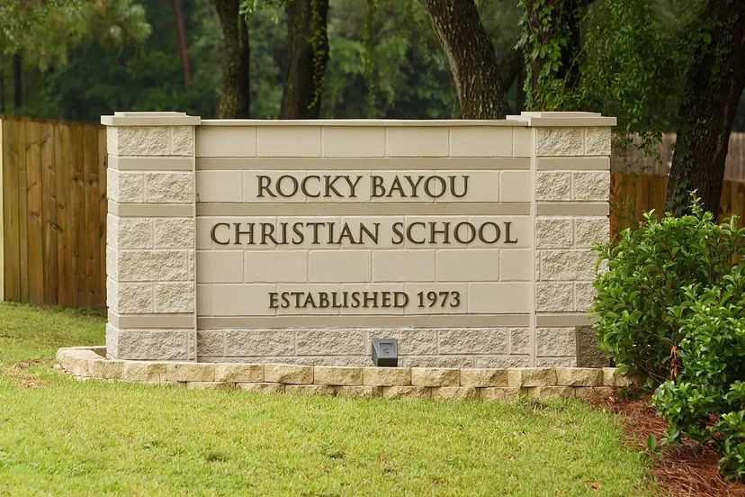 L'insegna in pietra mostra il nome Rocky Bayou Christian High School e l'anno di fondazione 1973 davanti a un fogliame verde.