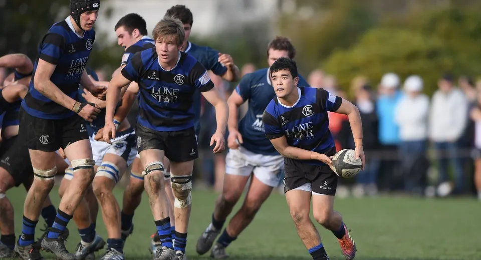 Un gruppo di giovani giocatori di rugby in maglia blu gioca sul campo sportivo della Taroona High School.