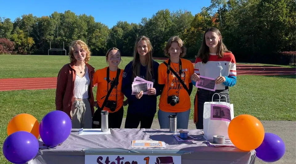 Un gruppo di studentesse della Acton High School si trova su un prato davanti agli alberi, tra palloncini colorati e decorazioni.