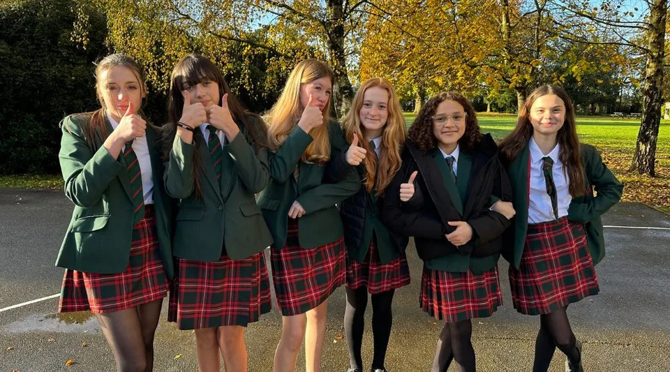 Un gruppo di studentesse della Akeley Wood School in divisa scolastica si trova insieme in un parco con alberi e piante.