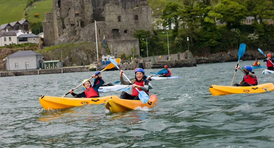 Studenti del Templemichael College pagaiano in kayak colorati su uno specchio d'acqua con un castello storico sullo sfondo.