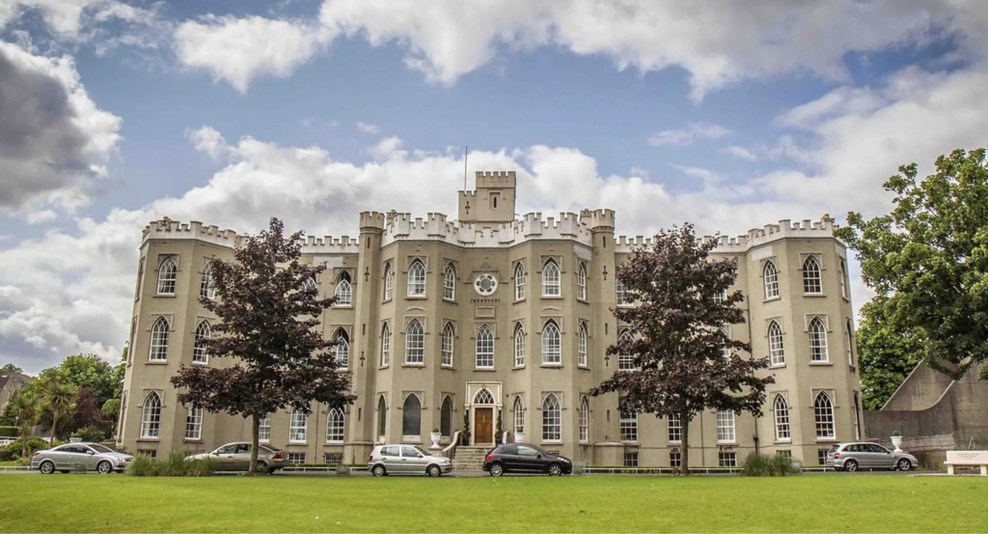 L'edificio storico del Blackrock College con facciata a merli, un prato verde e auto parcheggiate.