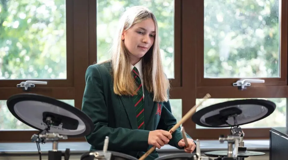 Una studentessa della Akeley Wood School suona la batteria in un accogliente ambiente interno con una finestra sullo sfondo.