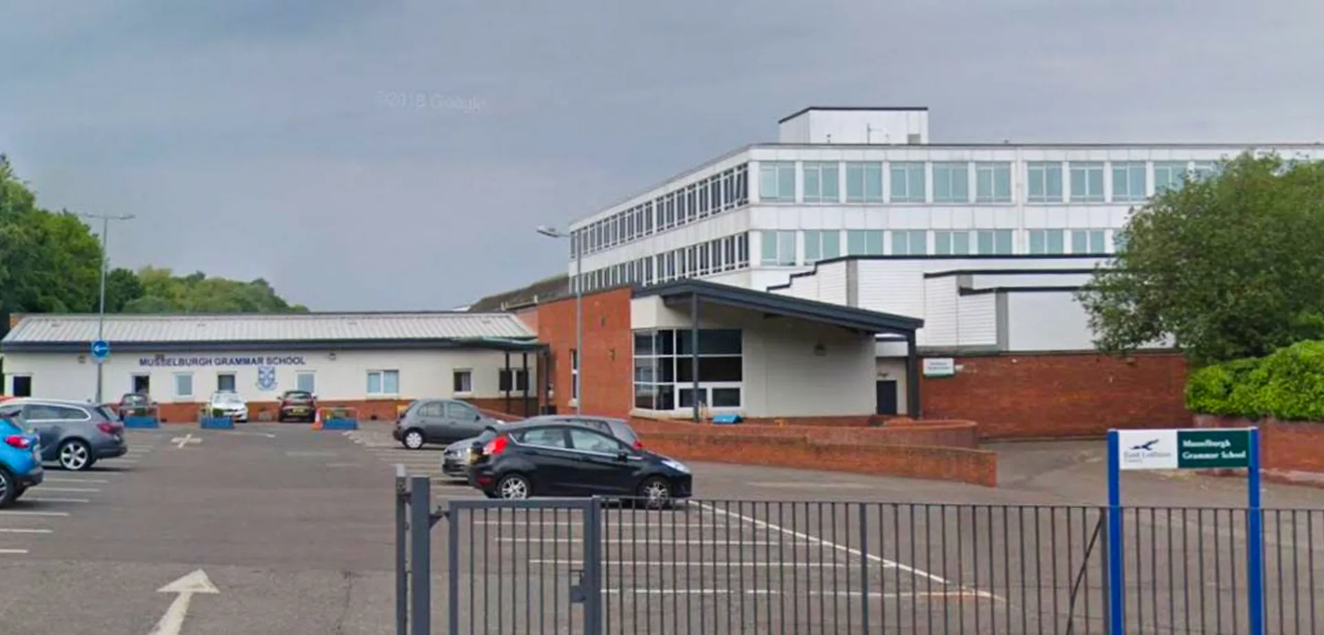 L'edificio a più piani della Musselburgh Grammar School con facciata in vetro e parcheggio in primo piano è circondato da alberi.