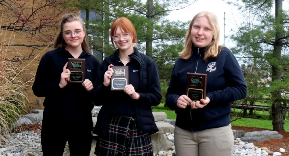 Tre studentesse della Mitchell District High School con dei premi in mano sono in piedi davanti a un muro di pietra e ad alcuni alberi.