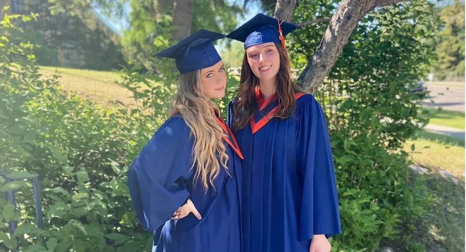 Due studentesse della James Fowler High School si trovano all'aperto nei loro abiti da diploma con alberi sullo sfondo.