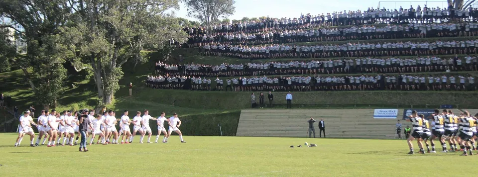 Un grande gruppo di studenti della New Plymouth Boys' High School si riunisce su un prato davanti a una struttura alta e densamente costruita.