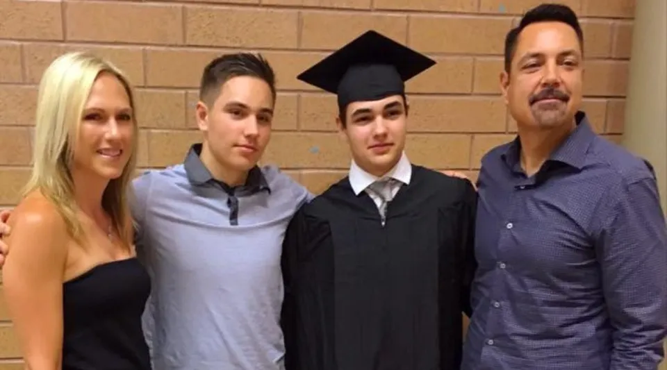 Un gruppo di quattro persone, tra cui uno studente con la toga da diploma, si trova davanti a una parete di mattoni nel terreno della Dr. Frank J. Hayden Secondary School.