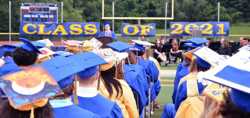 Un gruppo di diplomate e diplomati della Vernon Township High School indossa toghe blu e dorate davanti a uno striscione con la scritta Class of 2021 su un campo sportivo.